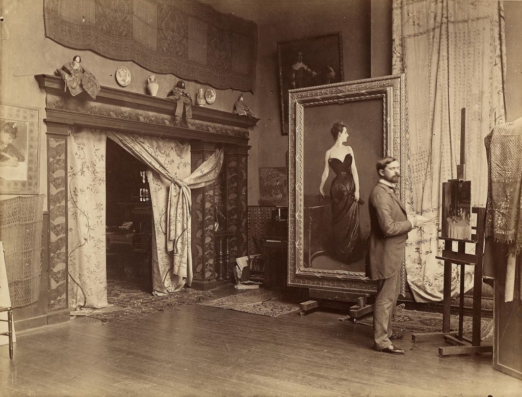 Sepia photograph, interior, many curtains and draperies over doors and windows. A man in a suit with a small beard and mustache stands in front of a large painting of a woman (the infamous Madame X portrait), while working on a smaller canvas to the side of it