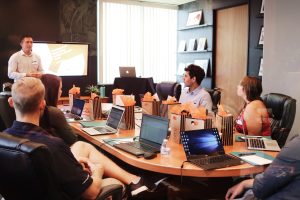 man standing in a conference room with 5 people sitting at a table