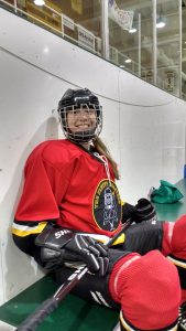 A young girl in her hockey uniform. She has long blonde hair in a ponytail.