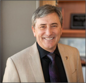 Image of one of the authors, Bob Boudreau. A smiling male with grey hair, wearing a black button up shirt with a purple tie and a tan suit jacket.