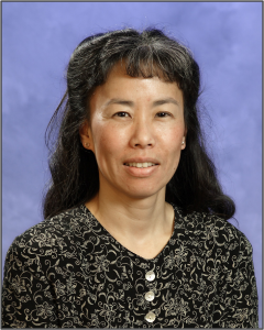 Picture of one of the co-authors, Rumi Graham. A smiling woman with dark hear wearing a dark shirt with light floral patterns against a blue background.