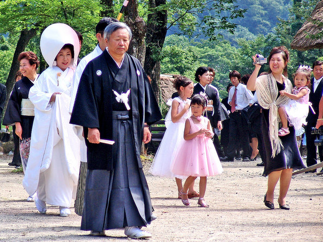 1.2.0 Descriptive image showing people outdoors at a traditional Japanese wedding. The bride and groom are walking behind a religious official, all dressed in traditional garb. Some attendees are dressed in kimono.