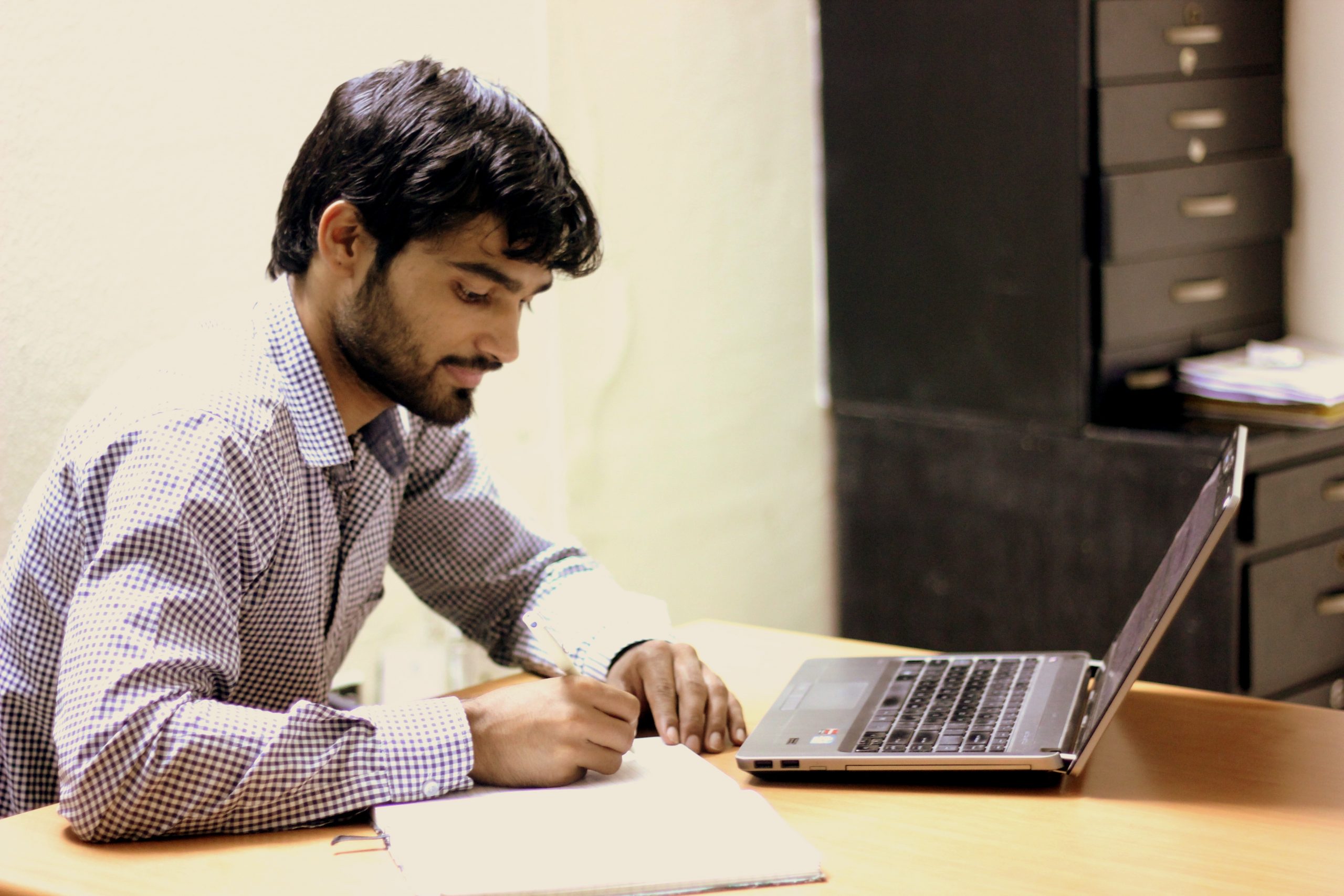 Descriptive image showing a man writing in a notebook while seated at a laptop.