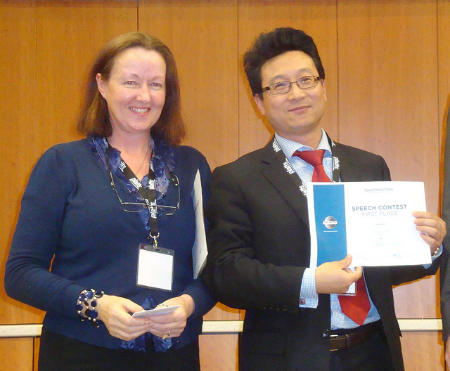 Descriptive image showing a woman standing next to a man; the man is smiling and holding up a Speech Contest certificate. (7.4.0)
