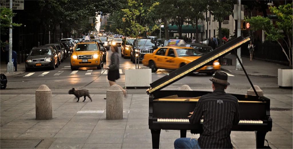A photograph shows a person playing a piano on the sidewalk near a busy intersection in a city.