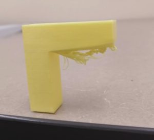 An upside down L-shaped 3D printed model. Yellow model on a grey background. An unpleasant droopy mess of filament hangs where the 3D printer attempted to print on an unsupported area.